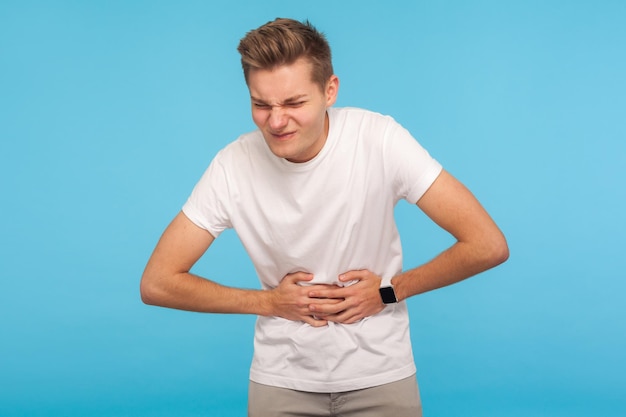 Sick man grimacing from stomach ache severe abdominal distress feeling discomfort in belly symptoms of constipation indigestion gastrointestinal disorder studio shot isolated on blue background