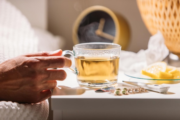 Photo sick man laying in bed under blanket and holding cup of hot tea with lemon