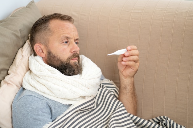 Sick man lying on sofa checking his temperature at home in the living room