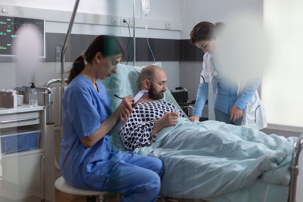 병동에서 클립 보드에 치료를 작성하는 동안 질병 증상을 설명하는 의료 간호사와 논의 침대에서 아픈 남자 환자 휴식