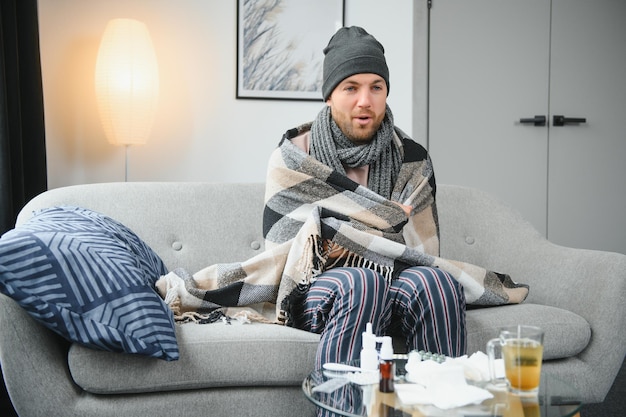 Больной мужчина сидит дома на сером диване с одеялом