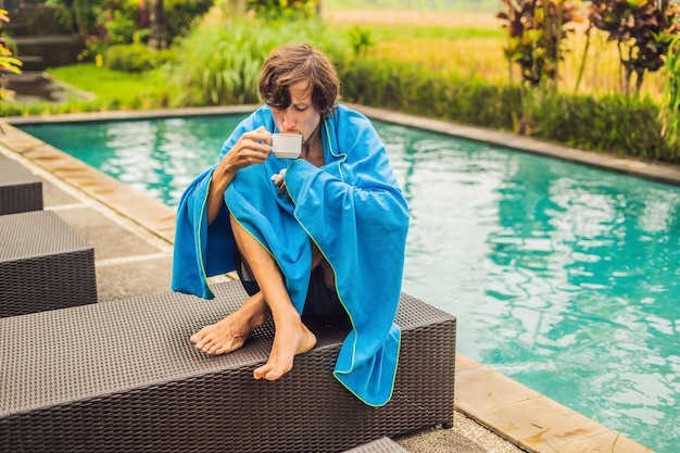 Sick man traveler The man caught a cold on vacation sits sad at the pool drinking tea and blows his nose into a napkin His son is healthy and swimming in the pool Travel insurance concept