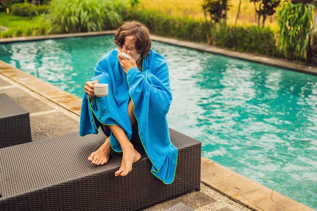 Sick man traveler The man caught a cold on vacation sits sad at the pool drinking tea and blows his nose into a napkin His son is healthy and swimming in the pool Travel insurance concept