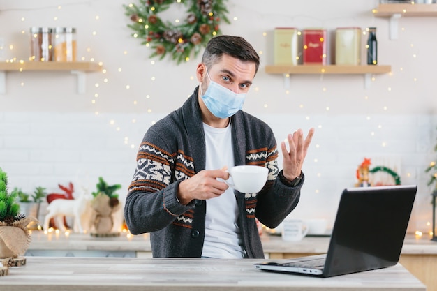 A sick man with a cold in a protective mask works remotely from home using a laptop during the New Year and Christmas holidays communicates with colleagues via video