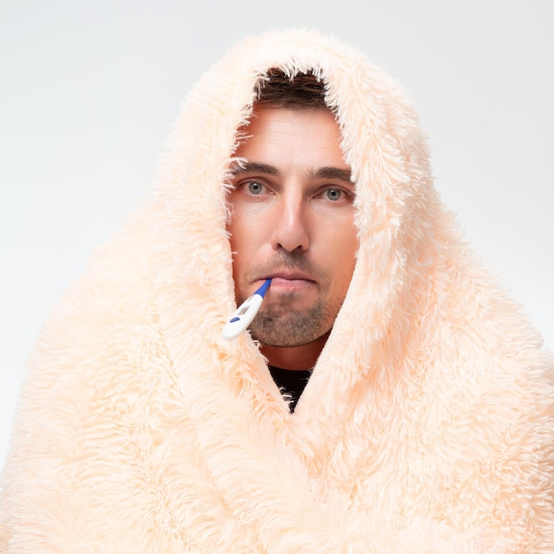 A sick man wrapped in a blanket holds a thermometer in his mouth