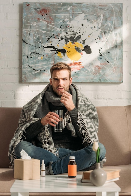 Sick man wrapped in blanket sitting on sofa and taking medicines