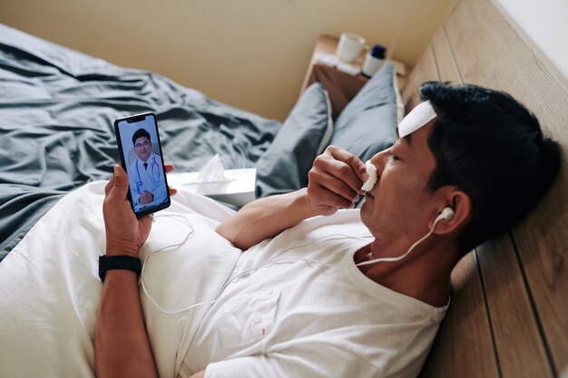 Sick mature Asian man blowing nose when video calling his doctor