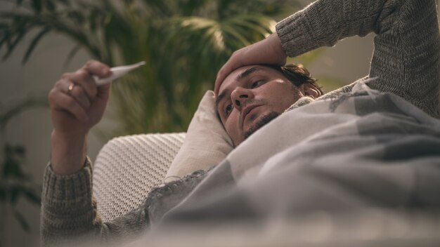 Sick men lying on a sofa and putting his hand on head, holding thermometer.