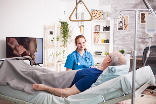 sick old man laying on bed in nursing home and talking with female nurse.