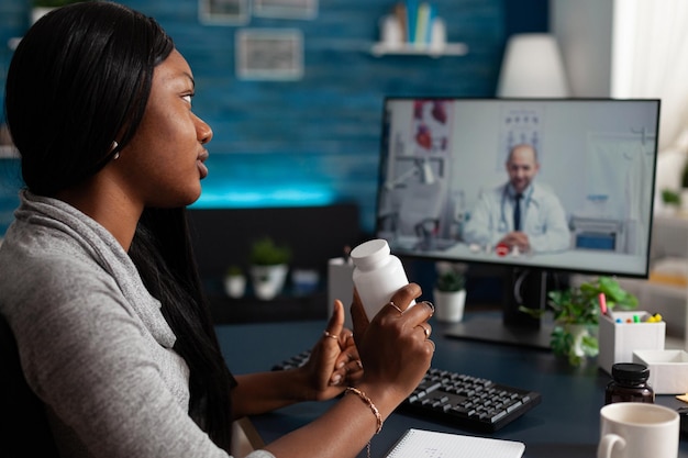 Больной пациент держит бутылку таблеток во время видеозвонка с медиком, делая удаленную онлайн-консультацию на компьютере. Женщина спрашивает врача о лечении на видеоконференции дома.
