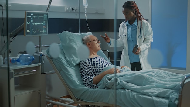 Sick patient laying in hospital ward bed at facility waiting\
for african american doctor to give bottle of pills as medical\
treatment. medic curing old man with health problems at clinic