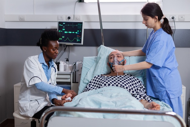 Sick patient lying in bed while woman nurse putting oxygen mask monitoring respiratory illness