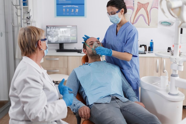 Sick patient sitting on dental chair during stomatology\
consultation while dentist nurse putting oxygen mask before teeth\
surgery in stomatological office room. medical team wearing face\
mask