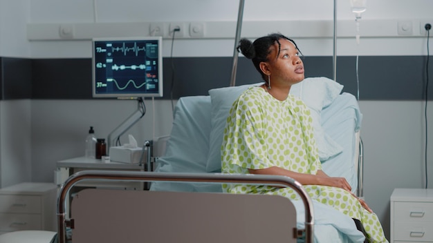 Sick patient waiting on doctor to give treatment and support\
while sitting in hospital ward bed. woman with disease receiving\
medical care at clinic with heart rate monitor and iv drip bag