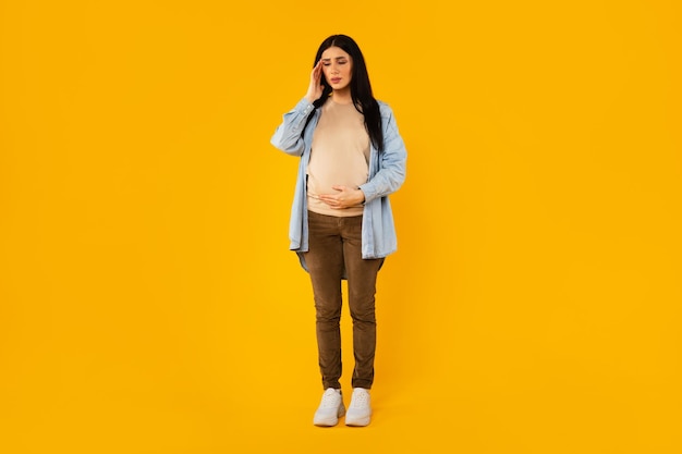 Sick pregnant woman suffering from headache standing isolated over yellow background full length shot