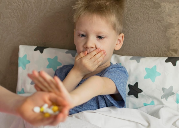 A sick sad child with a temperature and a headache lies in bed\
refuses to drink medicine and pills flu colds disease virus