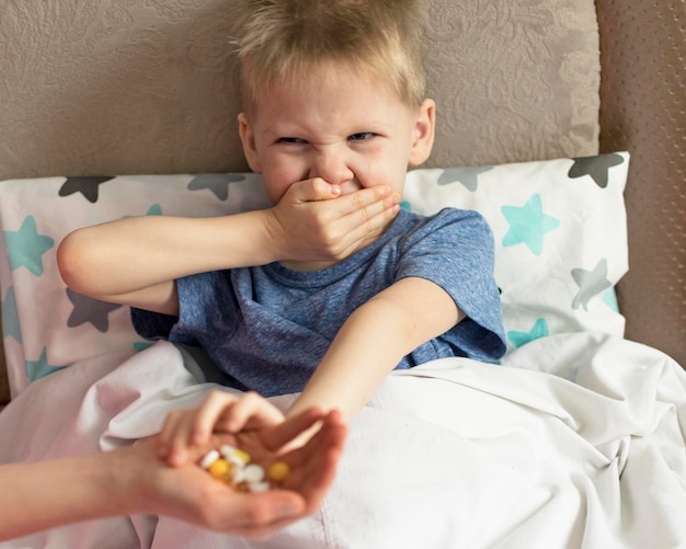 A sick sad child with a temperature and a headache lies in bed refuses to drink medicine and pills Flu colds disease virus