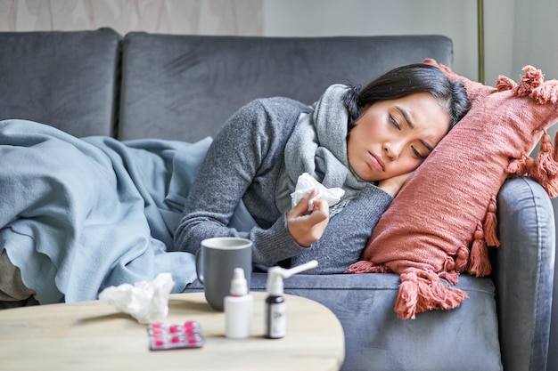 Sick sad korean woman lying on sofa feeling unwell catching cold flu and temperature looking upset t