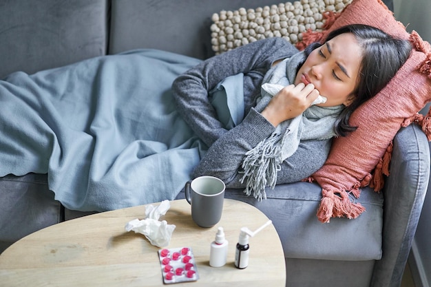 Sick sad korean woman lying on sofa feeling unwell catching\
cold flu and temperature looking upset taking medication