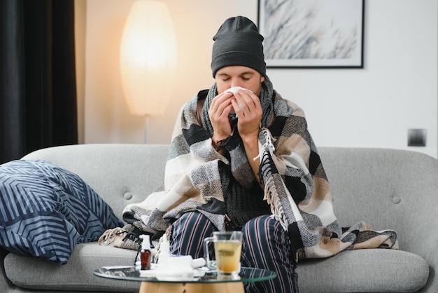 Sick sad man sits on couch at home suffers from runny nose flu disease coronavirus pandemic covid epidemic sneezes Unwell guy feeling bad fever virus illness symptoms indoor