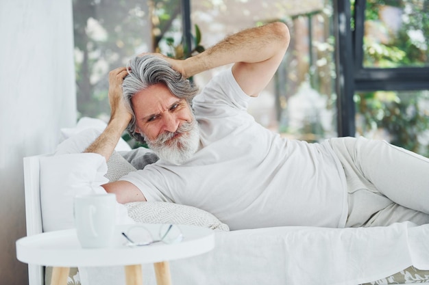 Sick senior stylish modern man with grey hair and beard indoors lying on the bed and feels bad