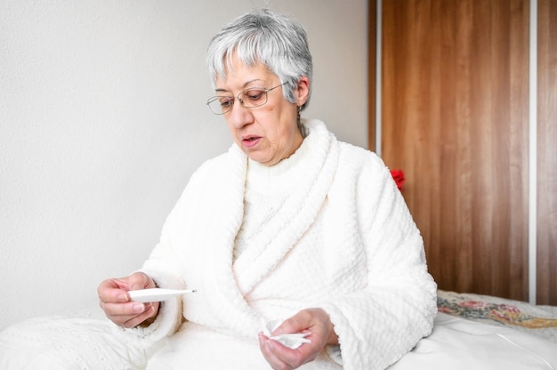 Sick senior woman holding tissue and thermometer sit on bed.