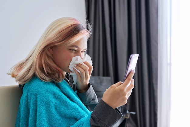 Sick teenager sneezing in handkerchief with symptoms of viral disease makes video call to doctor. Girl sitting at home receiving online consultation using webcam on smartphone