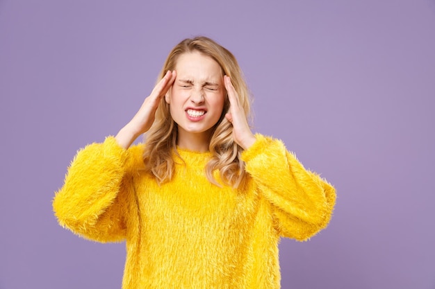 Sick tired young woman girl in yellow fur sweater posing isolated on pastel violet background. People lifestyle concept. Mock up copy space. Keeping eyes closed, put hands on head, having headache.