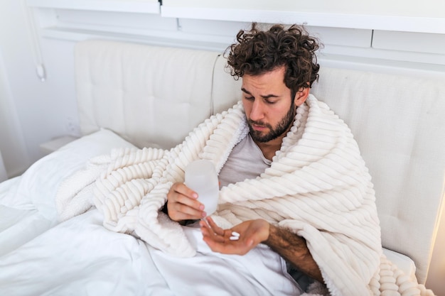健康管理の概念で薬の錠剤を持っている寒さと冬のインフルエンザウイルスに苦しんでいるパジャマを身に着けているベッドに横たわっている病気の無駄な男