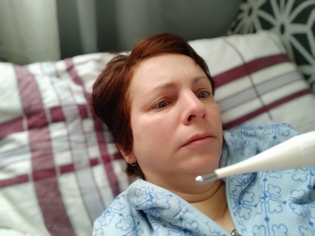 Photo sick woman in bed headache runny nose and cold paleness of the face and faintness of the body feeling unwell the patient lies on a pillow holds a thermometer and measures the temperature