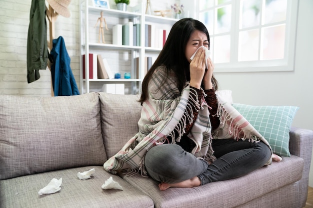 Sick woman covered with blanket sitting on couch. asian illness
lady with seasonal infections flu allergy and runny nose using
tissue napkins a lot on sofa at home. young female watch tv
sneezing.