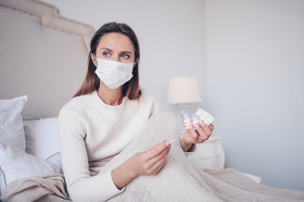 Sick woman in face protection mask lying in bed holding thermometer and pills at home quarantine isolation.