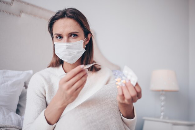 Sick woman in face protection mask lying in bed holding thermometer and pills at home quarantine isolation.