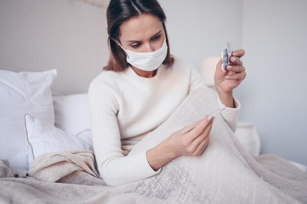 Sick woman in face protection mask lying in bed holding thermometer and pills at home quarantine