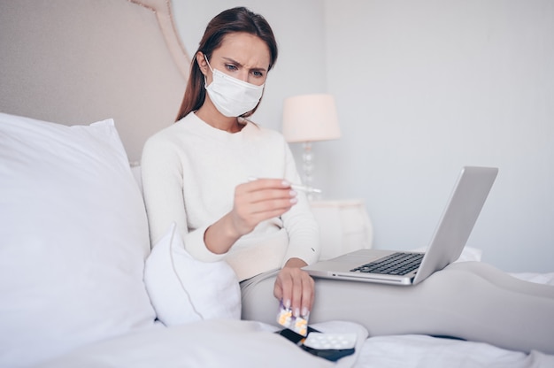 Sick woman in face protection mask lying in bed with laptop holding thermometer and pills at home