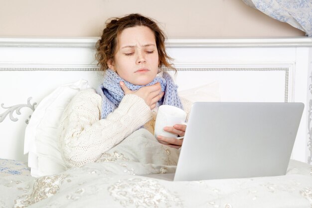 아픈 여자는 집에 있는 침대에서 노트북과 인터넷으로 기분이 나쁘고 쉬고 일합니다.