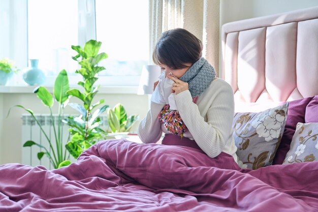 照片生病的女人拿着手帕打喷嚏擦拭她的鼻子坐在家中的床上健康问题感冒流感季节人们生活方式的概念