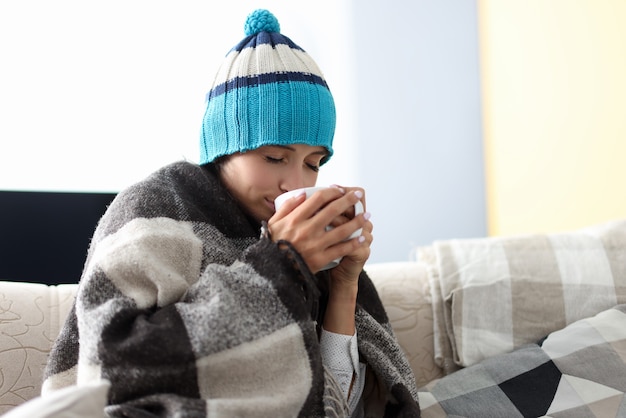 Фото Больная женщина в шляпе под одеялом держит чашку с лекарством