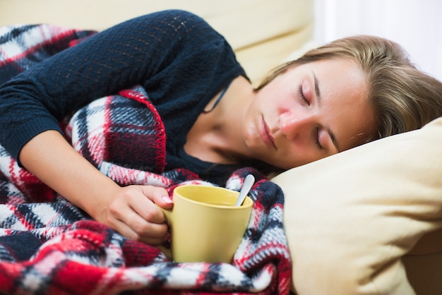 Sick woman lying on sofa under wool blanket with cup of tea