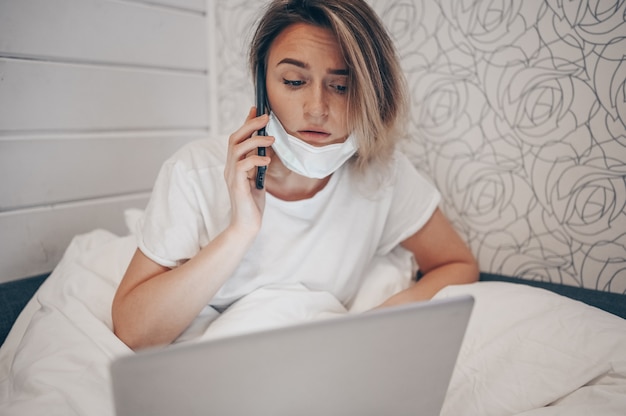 Donna malata in maschera protettiva sdraiato nel letto, parlando di smartphone e lavorando su un computer portatile durante l'isolamento di quarantena a casa covid-19 pandemia virus corona. distanza dal lavoro online dal concetto di casa.