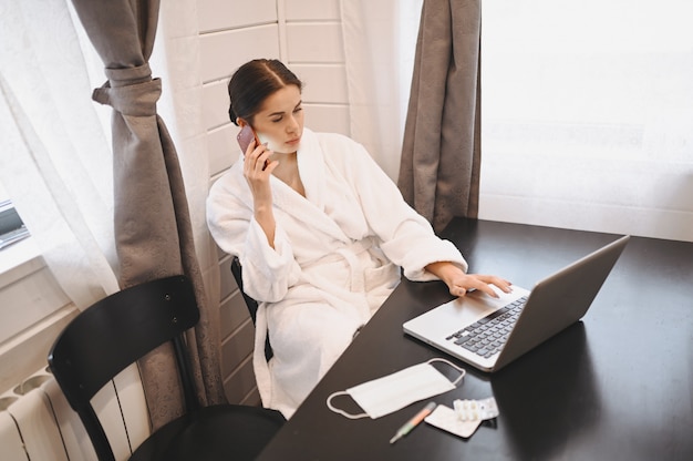 Sick woman speaking smartphone and working on a laptop during home quarantine isolation Covid-19 pandemic Corona virus. Distance work from home concept. Thermometer, pills and a medical mask on table