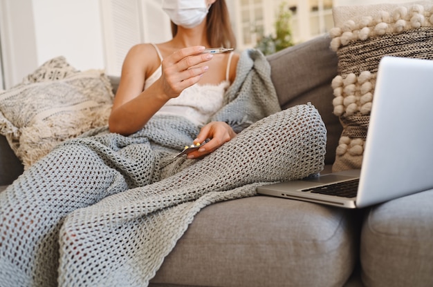 Donna malata che indossa la maschera di protezione del viso, seduto sul divano con il computer portatile e tenendo termometro e pillole durante l'autoisolamento di quarantena domestica.