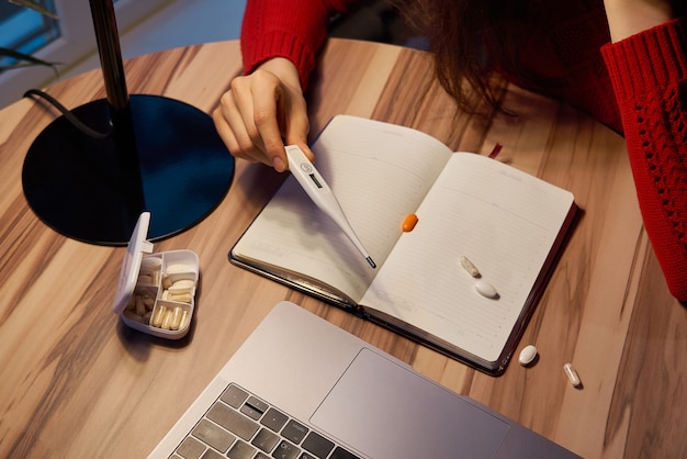 больная женщина с таблетками и термометром на столе работает на ноутбуке из дома на карантин изоляции.