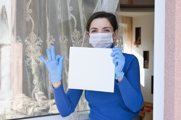 Sick woman with virus protection face mask and gloves looks out of the window