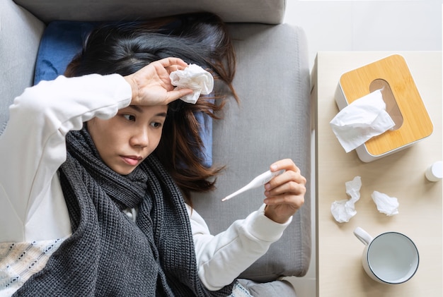 사진 아픈 젊은 아시아 여성이 집에서 소파에 누워 추위를 느낍니다. 온도계와 이마를 만지는 손으로 체온을 측정하는 소녀. 겨울 감기와 독감 개념입니다. 평면도.