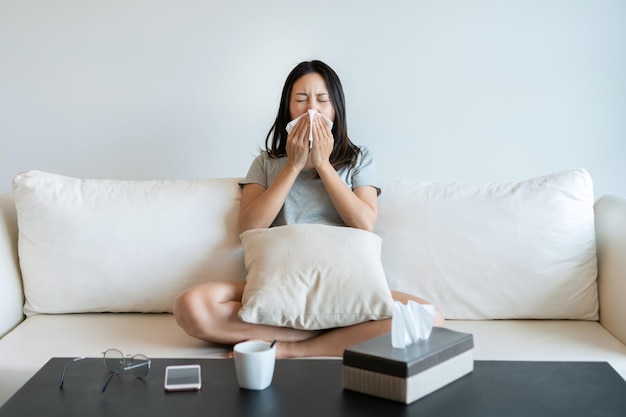 Sick young Asian woman sitting on couch blowing her nose on a tissue conceptual of healthcare seasonal flu rhinitis or an allergic reaction in hay fever