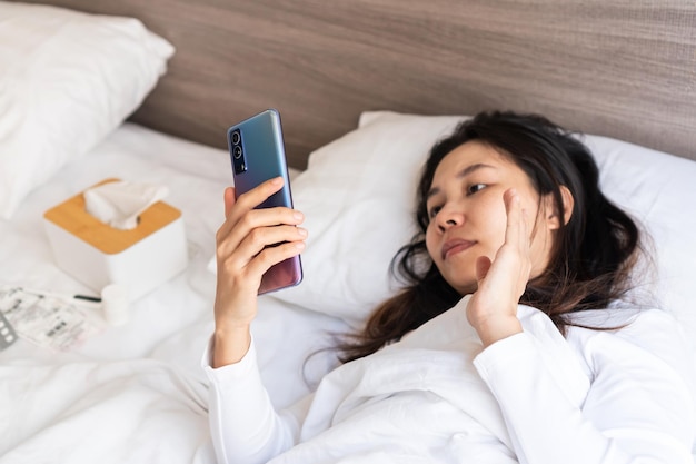 La giovane donna asiatica malata dorme sul letto in camera da letto a casa e videochiama con il medico o la sua famiglia sul telefono cellulare isolamento domestico di influenza fredda durante il concetto di pandemia di coronavirus primo piano