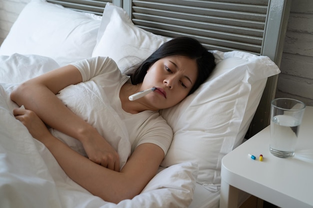 입 안의 체온을 측정하기 위해 온도계를 들고 침대에 누워 있는 아픈 젊은 여성. 여자 감기 발열 의료 건강을 잡아. 따뜻한 담요 커버 약 알약과 물 옆에 아픈 여자.