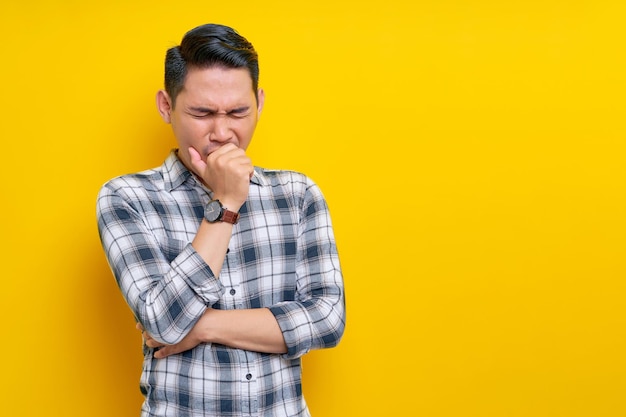 Sick young handsome Asian man 20s wearing a plaid shirt coughing covering mouth with hand keeping eyes closed isolated on yellow background People lifestyle concept