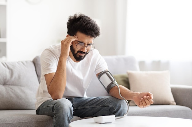 Sick young indian man checking blood pressure with modern tonometer at home